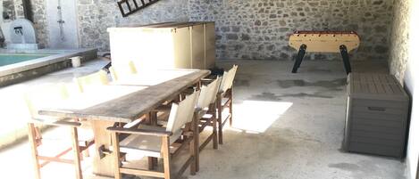 A stone-walled room with a long wooden table and chairs, a small pool, and a wooden bench.