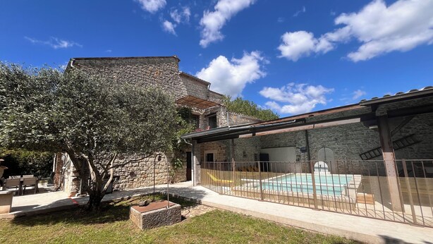 Un edificio de piedra con área de piscina, rodeado por una terraza cercada y un árbol.