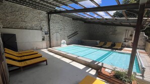 Un área de piscina al aire libre techada, con sillas de descanso, un muro de piedra y una estructura de techo metálico.
