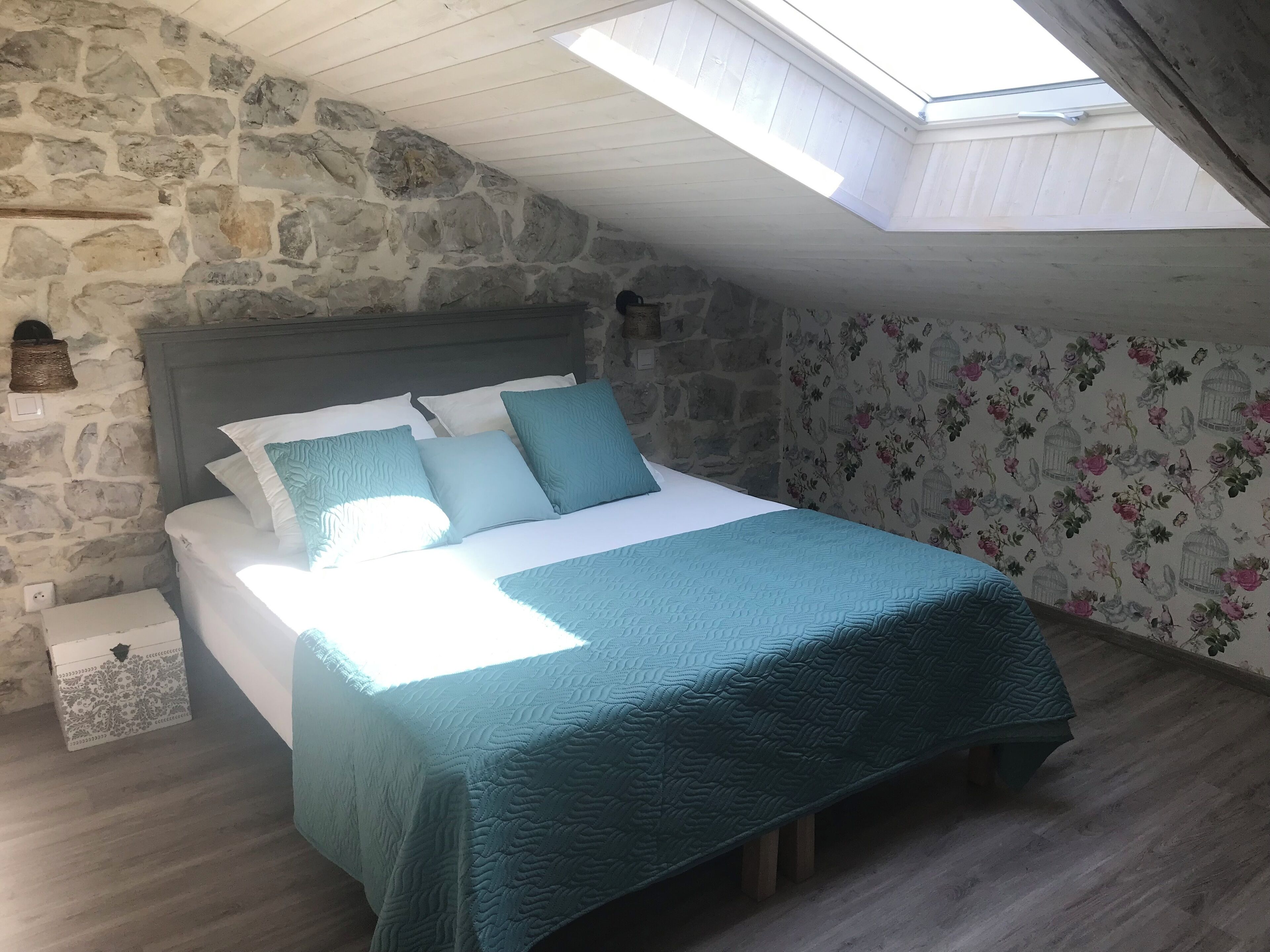 Ein Schlafzimmer mit Steinwand, Oberlicht und einem Bett mit türkisfarbenem Bettzeug sowie floralem Wandpapier.