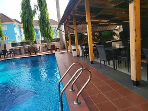 A swimming pool with a tiled deck and metal railings, surrounded by outdoor seating and a covered area with wooden beams. - Parklane Hotels  (Abuja)