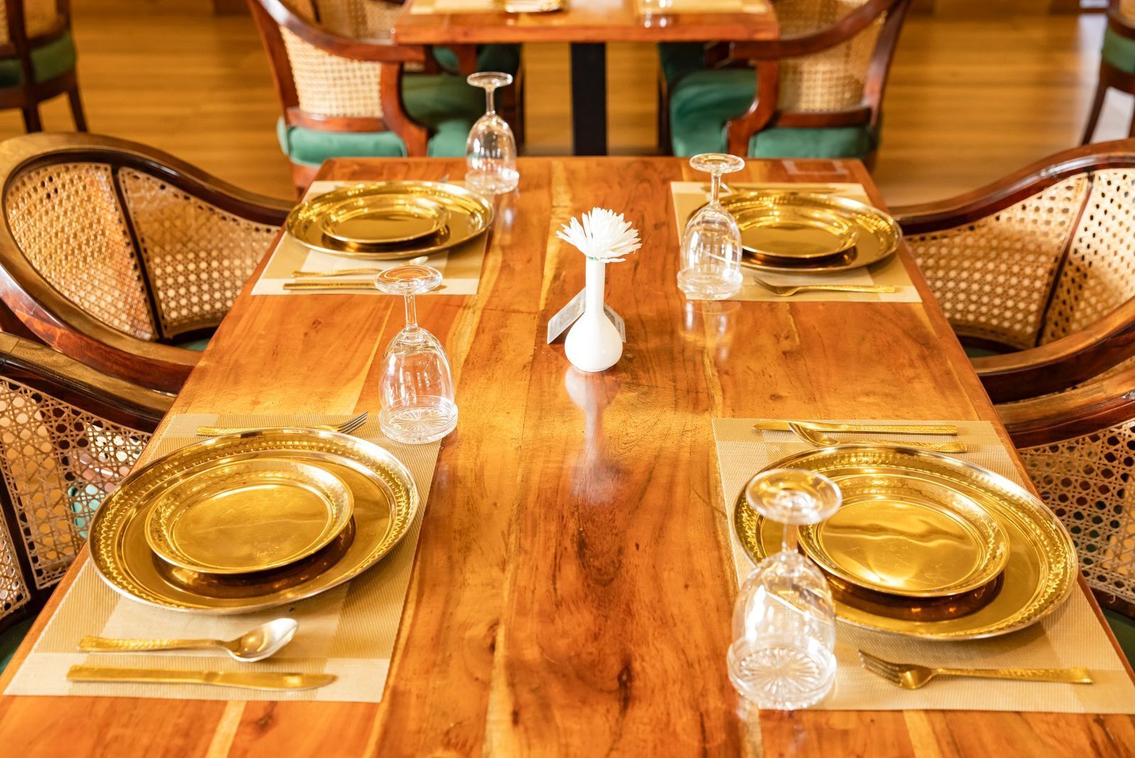 A dining table set with gold plates, cutlery, and glassware, surrounded by wicker chairs.