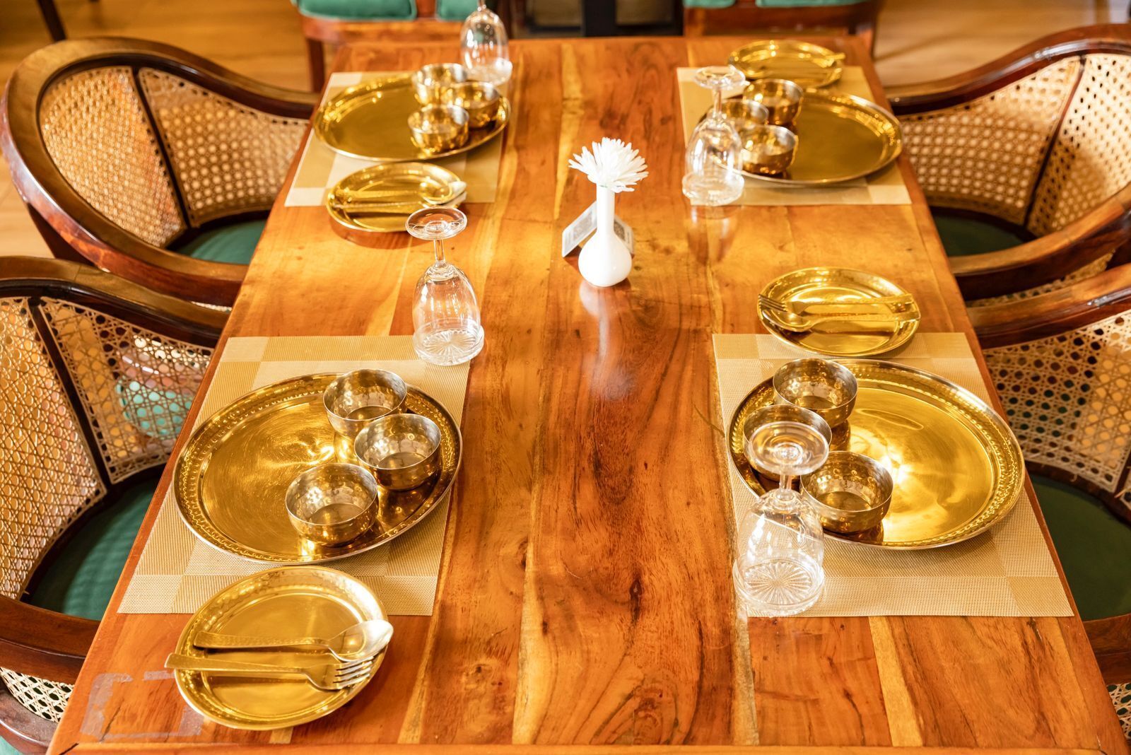 Mesa de jantar de madeira posta com pratos, tigelas e copos dourados, rodeada por cadeiras estofadas.