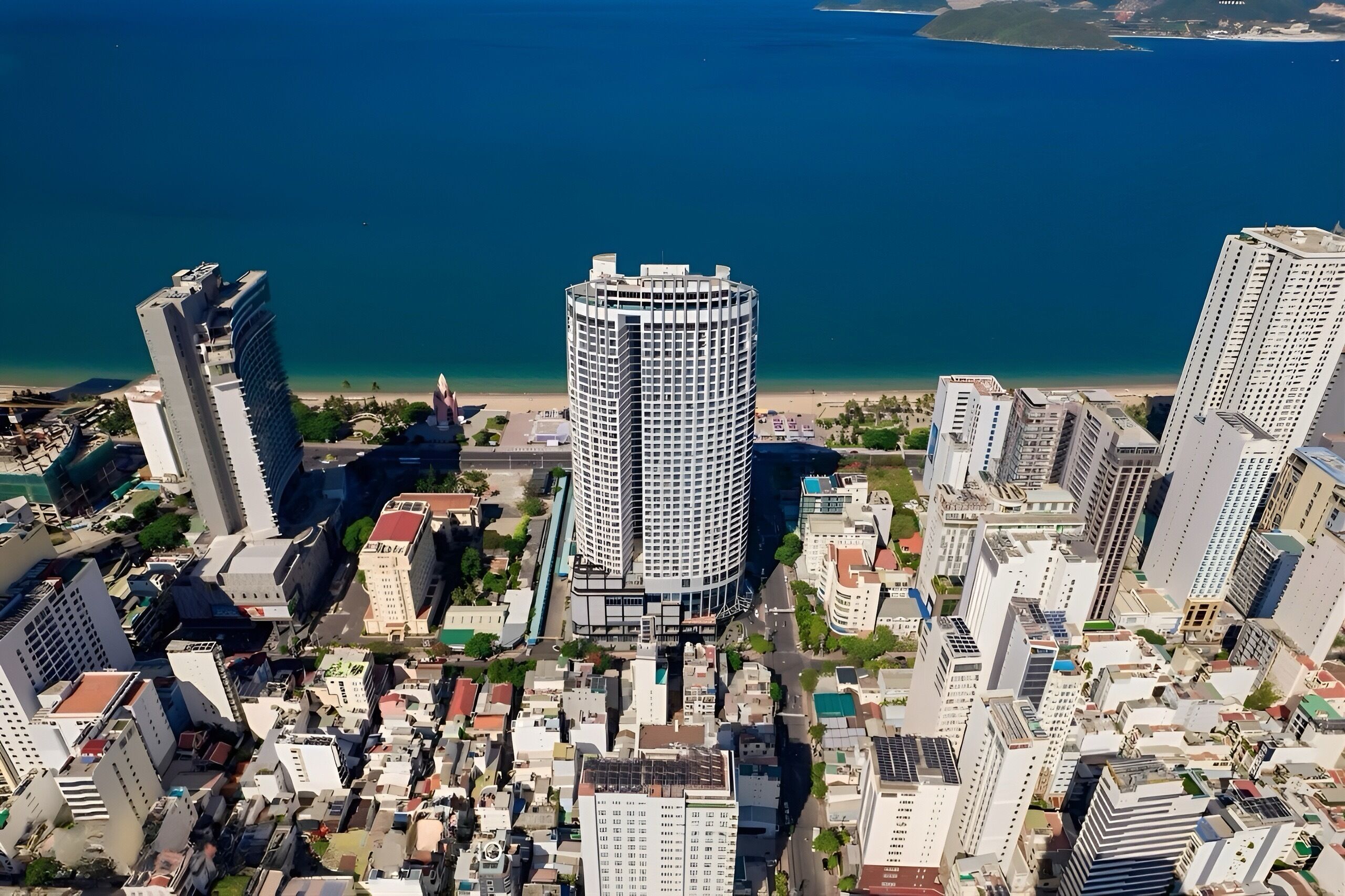 A high-rise building with a circular design, surrounded by other tall buildings, near a coastline.