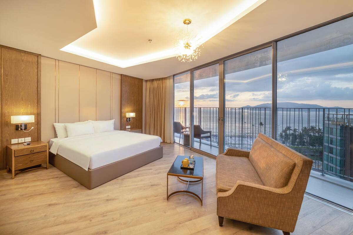 Quarto de hotel moderno com uma cama grande, um sofá, uma mesa pequena e uma varanda vista para a cidade.