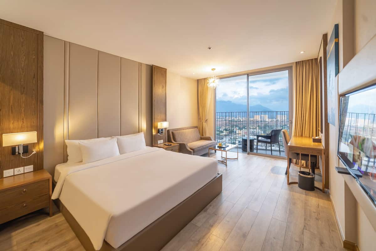 Quarto de hotel moderno com uma cama grande, escrivaninha, cadeira e varanda com vista para a cidade.