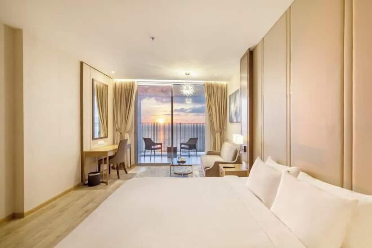 Quarto de hotel com uma cama grande, uma escrivaninha, uma cadeira e vista para o pôr do sol pelas portas da varanda.