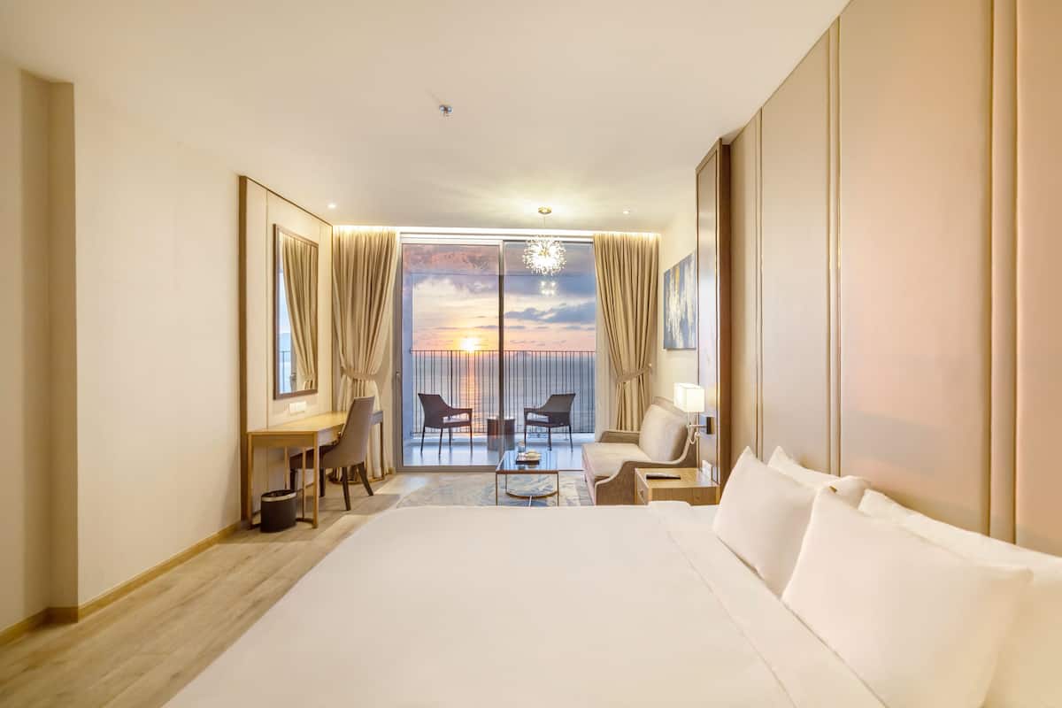 Quarto de hotel com uma cama grande, uma escrivaninha, uma cadeira, um sofá e varanda com vista para o pôr do sol.