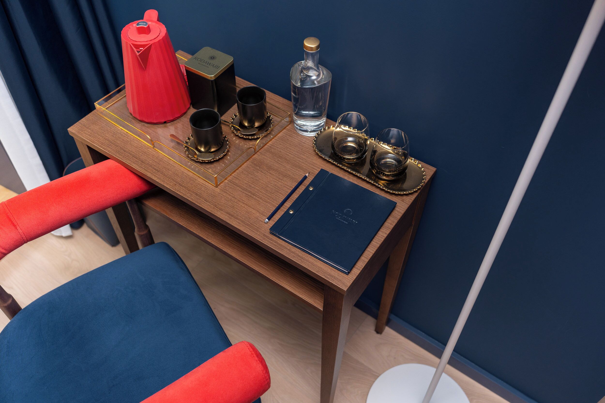 A wooden desk with a blue and red chair, a red kettle, a bottle of water, and a black notebook.