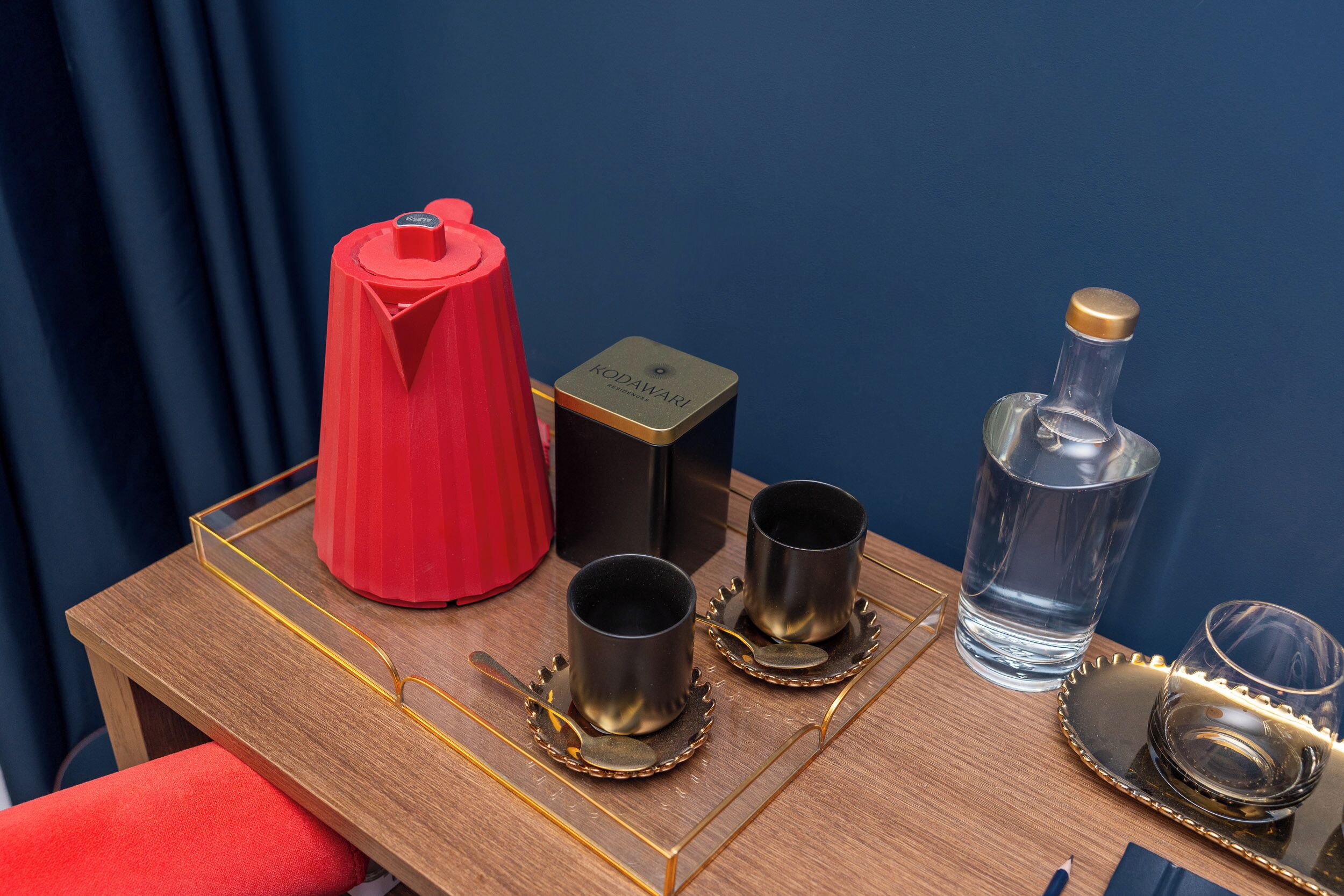 Una mesa de madera con un hervidor rojo, dos tazas negras, un vaso de agua, una botella y una cuchara.