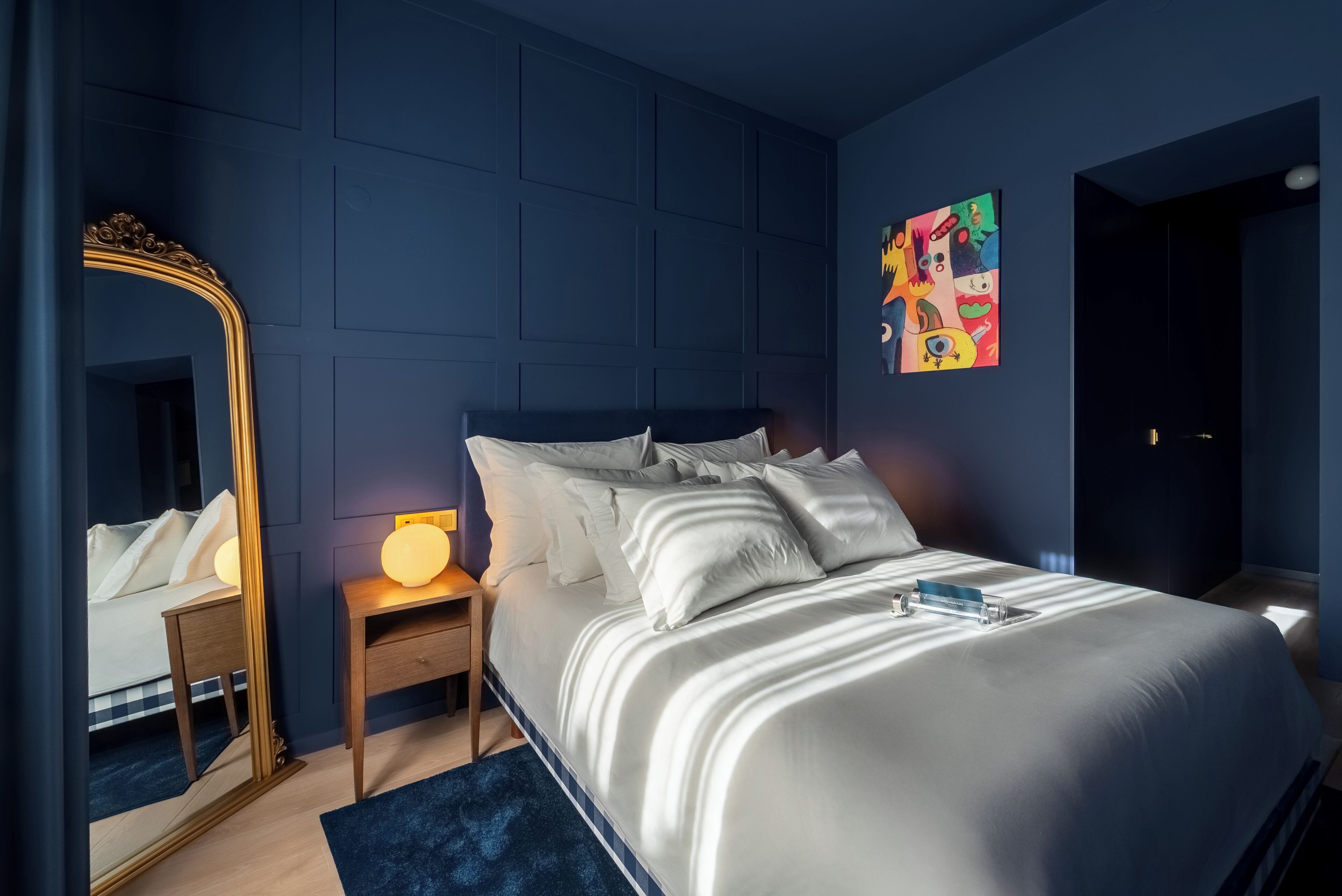 Un dormitorio con cama, mesita de noche, lámpara, espejo y un cuadro colorido en la pared.