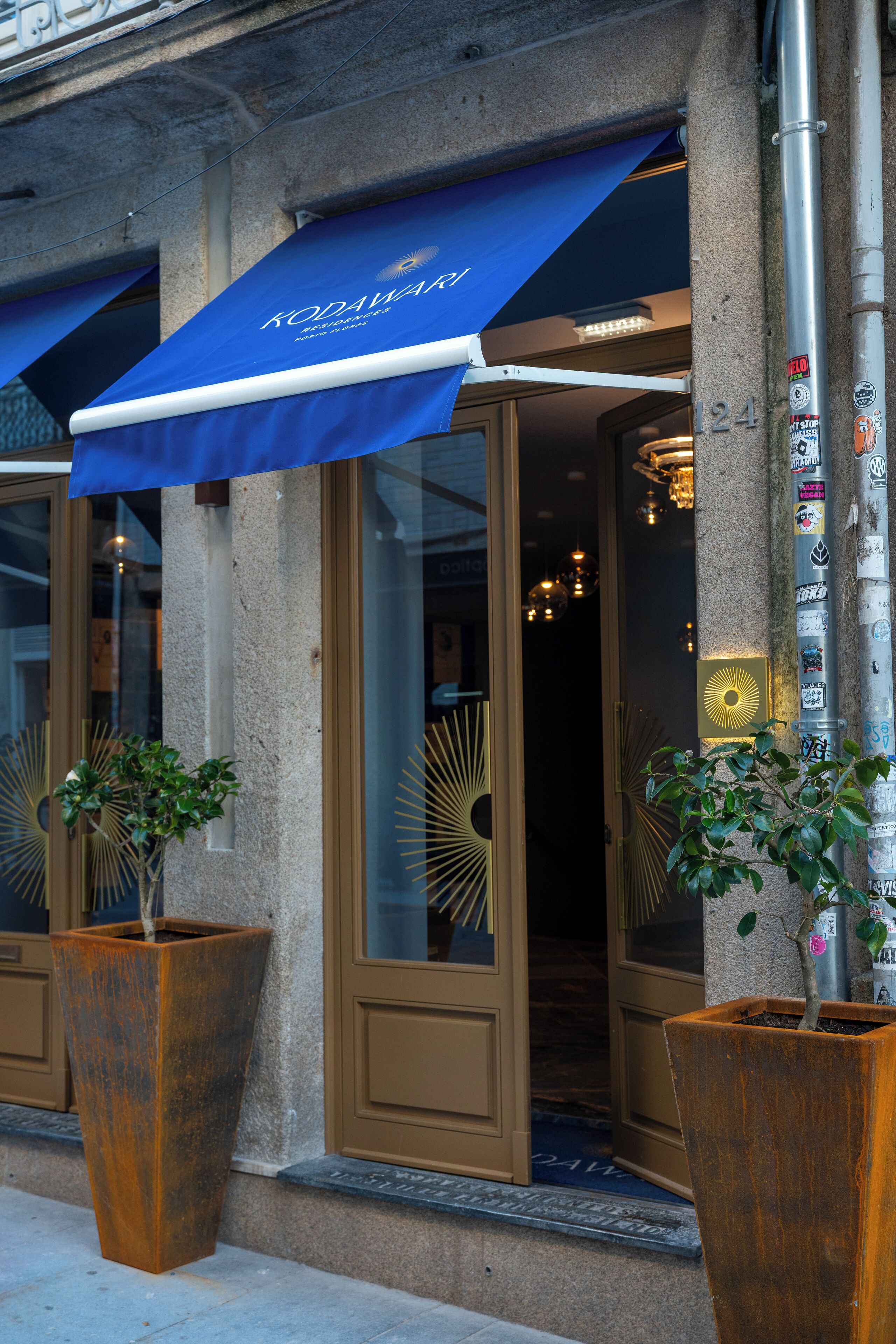A building entrance with a blue awning, large potted plants, and a door with glass panels.