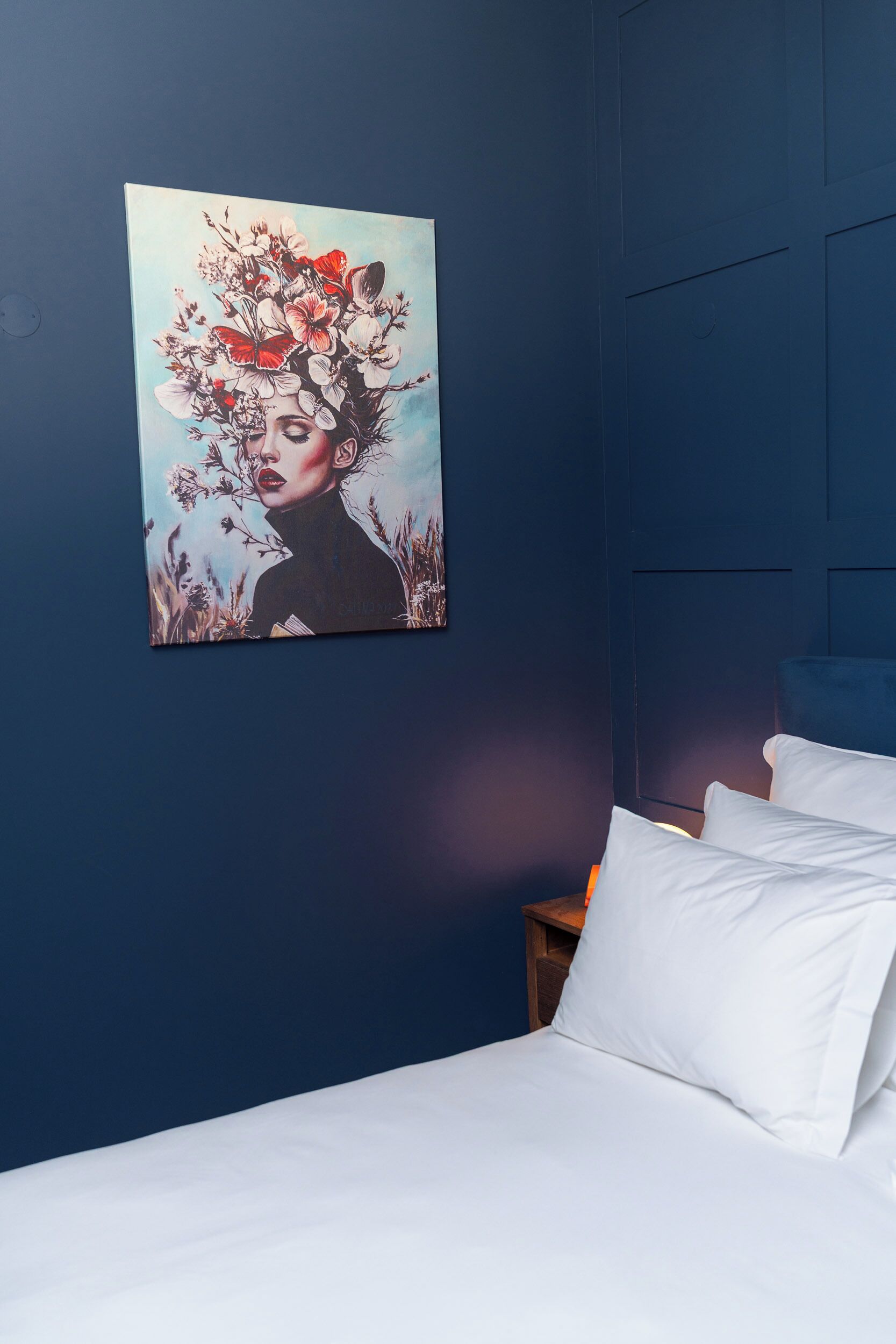 A bedroom with a blue wall, a white bed, and a painting of a woman with flowers.
