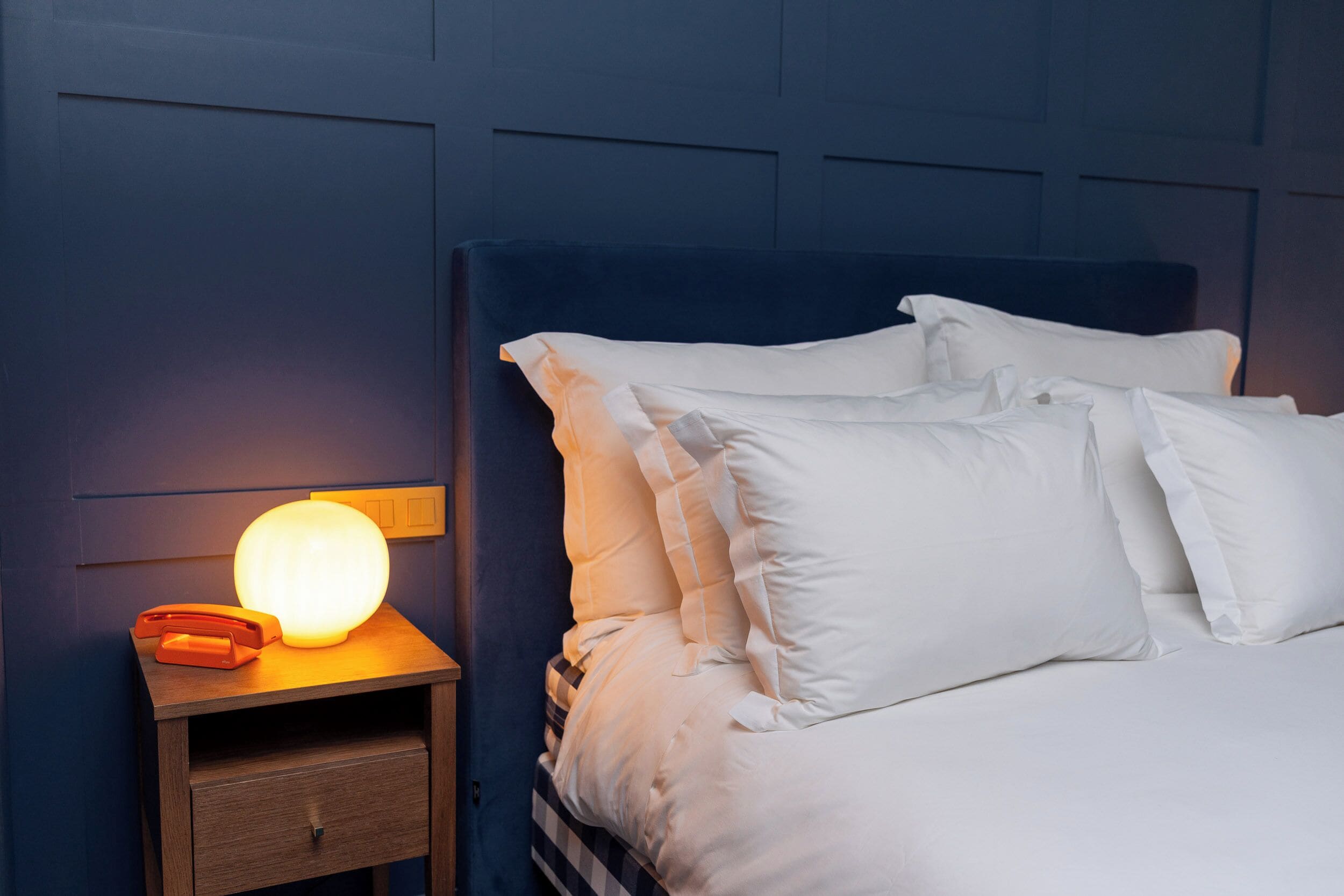 Una cama bien hecha con sábanas y almohadas blancas, una lámpara de noche y una mesita de noche de madera con un teléfono.