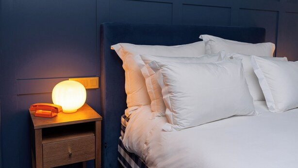 A neatly made bed with white linens and pillows, a bedside lamp, and a wooden nightstand with a telephone. - Hotel Kodawari Flores - By Kodawari Residences  (Porto)