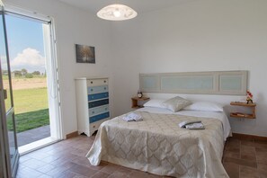 Un dormitorio con cama, cómoda, mesita de noche, un cuadro y vistas a través de un ventanal.