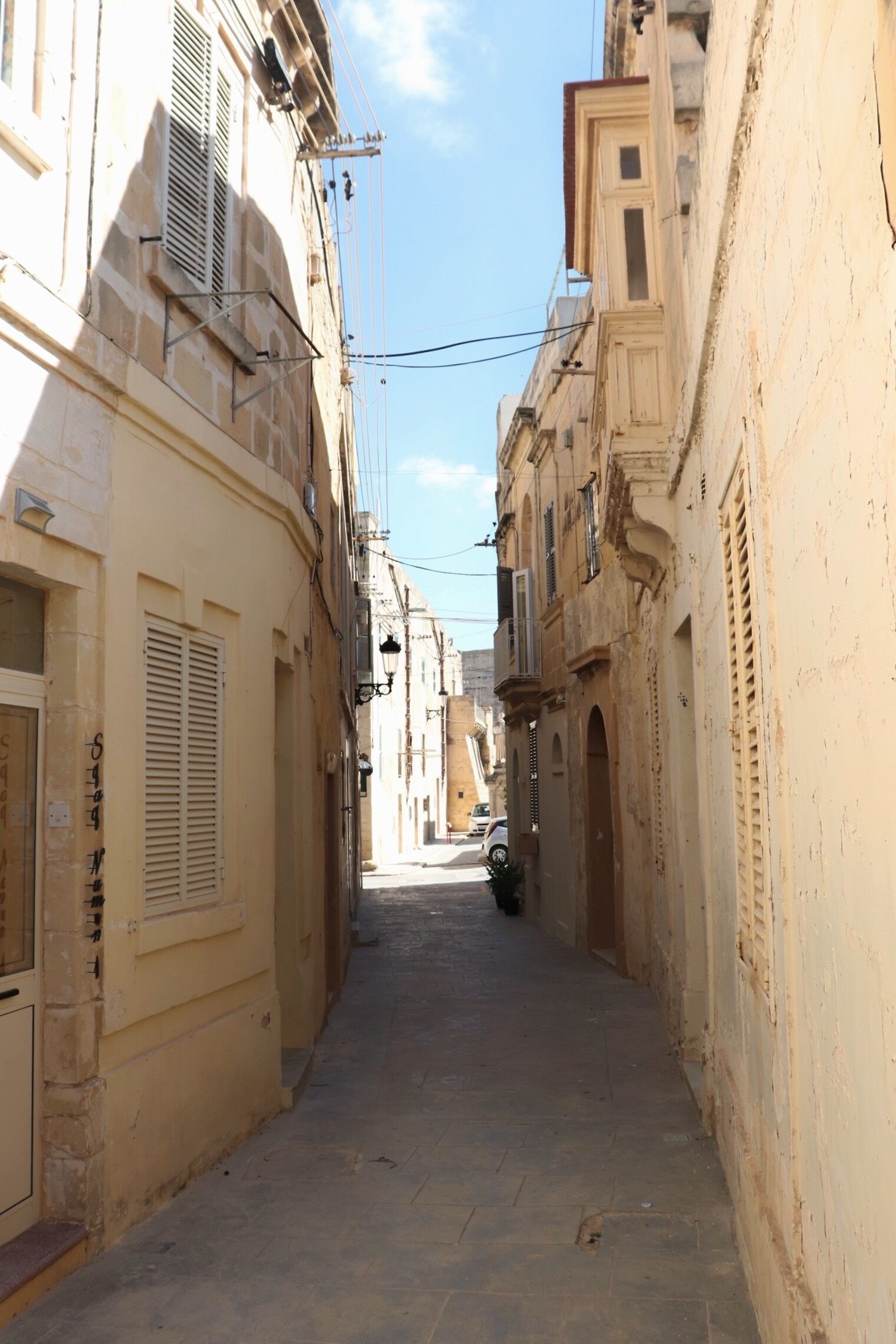 A narrow street with buildings on both sides, featuring balconies and shutters.