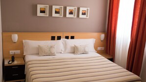 A hotel room with a large bed, two bedside tables, a nightstand, a wall-mounted lamp, and three framed pictures above the bed. - Hostal Albero by eme hoteles (Sanlucar la Mayor)