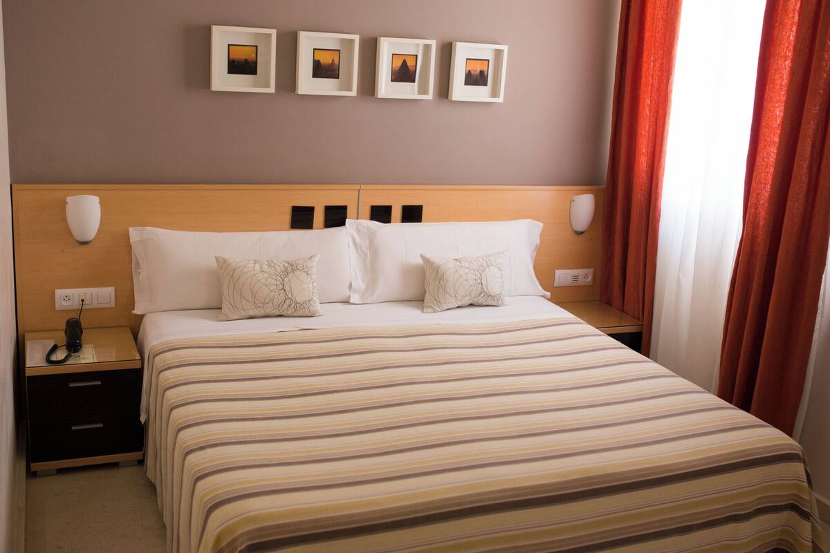 Une chambre d’hôtel avec un grand lit, deux tables de chevet, une petite table, une lampe fixée au mur et trois tableaux encadrés au-dessus du lit.
