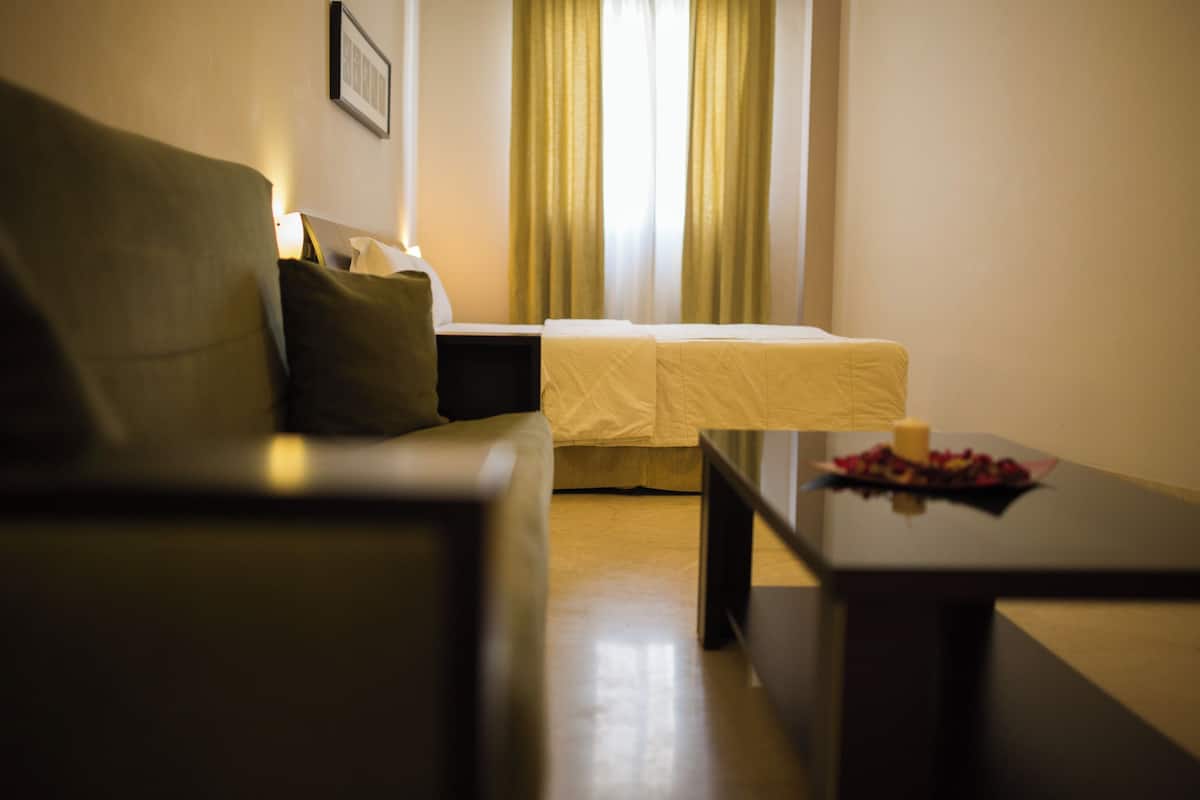 Une chambre d’hôtel avec un canapé, un lit recouvert d’une courtepointe blanche, une petite table avec une bougie et des fleurs, et une fenêtre avec des rideaux.