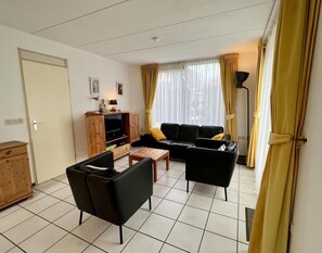 A living room with a black sofa, a wooden cabinet with a TV, and a lamp. - Holiday Bungalow With Wifi Near Beach (Sint Maartensvlotbrug)