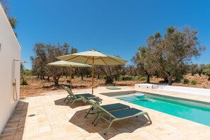 Un área de piscina con dos sillones de descanso y una sombrilla grande, rodeada de árboles y un edificio blanco.