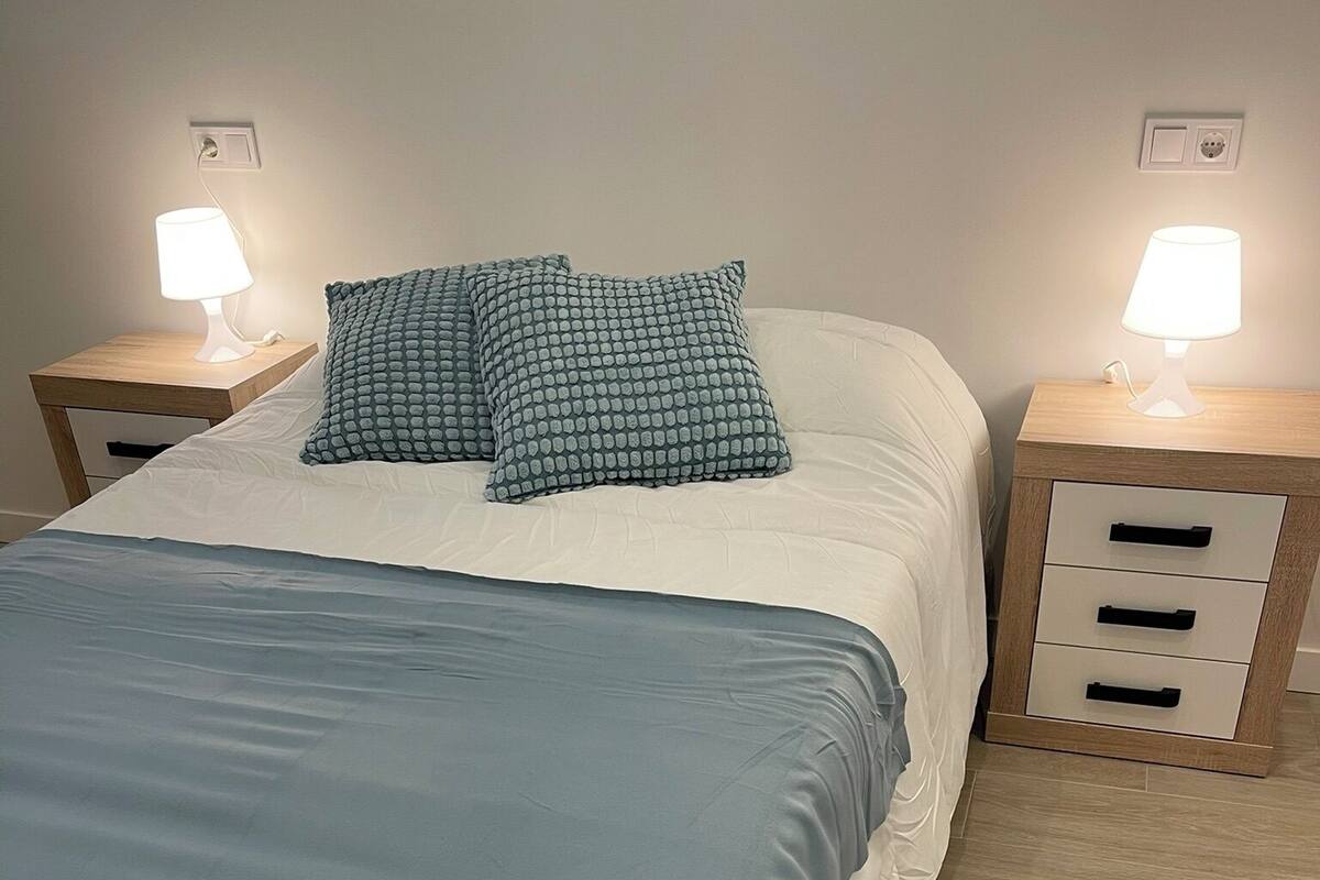 Un lit bien fait, recouvert d’une couverture bleue et agrémenté de deux coussins à motifs, avec de chaque côté des tables de chevet dotées de lampes et de tiroirs de rangement.