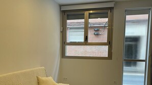 Una sala de estar moderna con un sofá marrón oscuro, dos cojines blancos, una mesita pequeña y un ventanal con vistas a un edificio de ladrillo.
