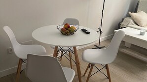 Un área de comedor con una mesa redonda, cuatro sillas blancas, un cuenco de fruta, un control remoto y una lámpara de pie.
