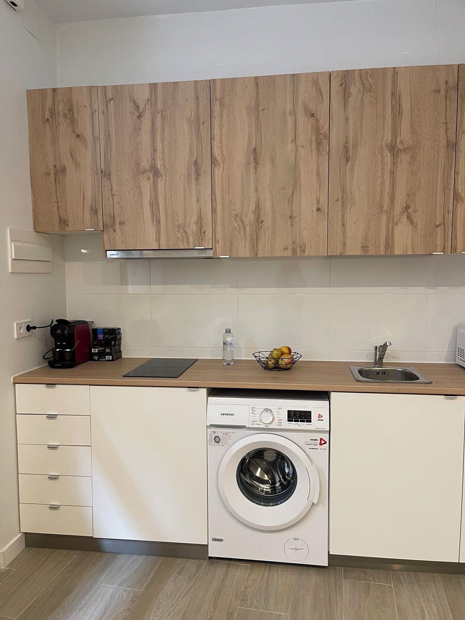 A compact kitchen with wooden cabinets, a built-in washing machine, a coffee maker, and a fruit bowl.