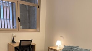 Un dormitorio con cama, escritorio y silla, mesita de noche con lámpara, y ventana con persianas.