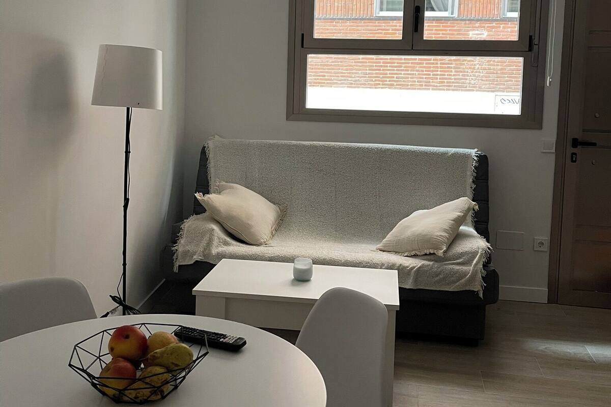 Un salon moderne avec un canapé, une petite table et une lampe. Il y a une fenêtre avec des stores et une vue sur un bâtiment à l’extérieur.