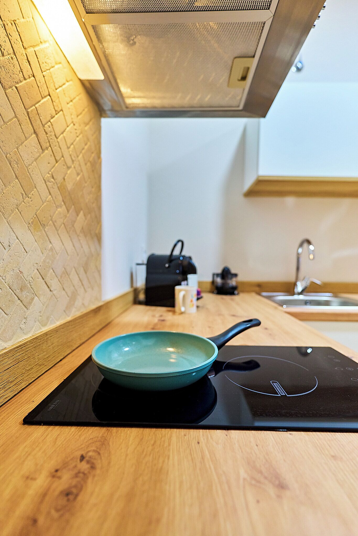 A kitchen with a brick wall, a wooden countertop, a black induction cooktop, a teal pan, a coffee maker, and a kettle.