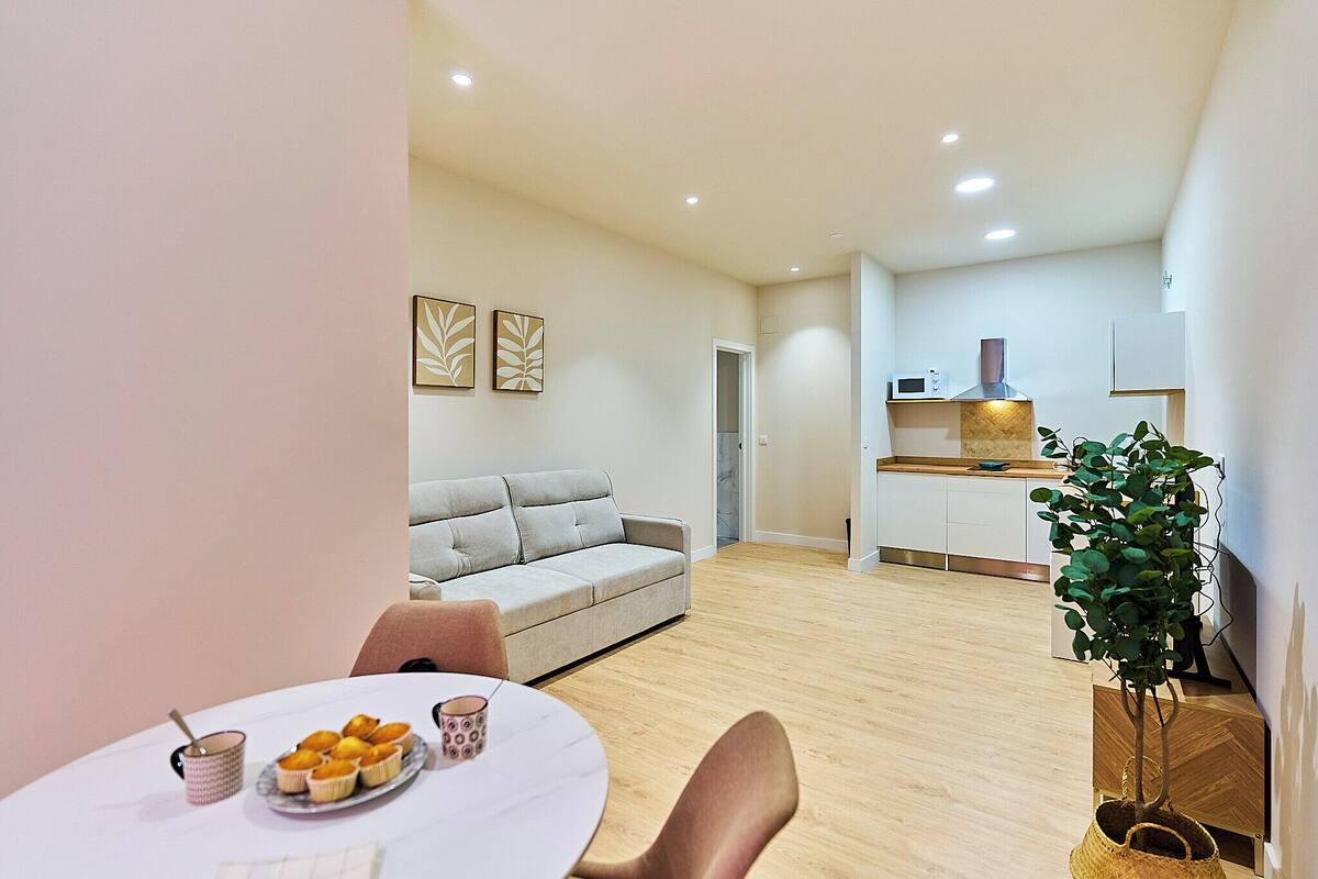 Una sala de estar moderna con un sofá, una mesa de comedor con muffins, y una cocina con una planta y armarios.