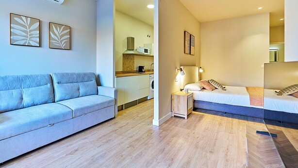 Una habitación de hotel moderna con sofá, cama, mesita de noche con lámpara y vista al área de la cocina.