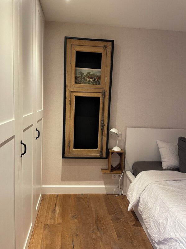 A bedroom with a wooden-framed picture, a bedside table with a lamp, a bed with white bedding, and a wooden floor. - Le petit Second, charming gîte for 2 people in the center of the town of Spa (Spa)