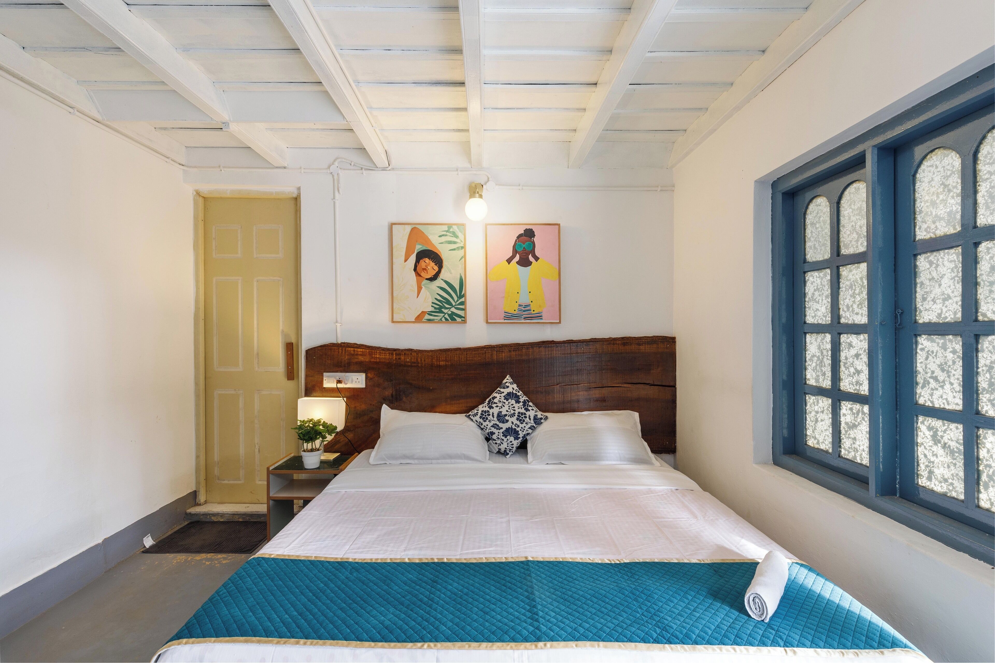 A neatly made bed with a blue quilt, a wooden headboard, and a window with a view.