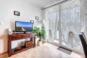 A living room with a TV, wooden entertainment unit, potted plants, and a view through a window with sheer curtains.