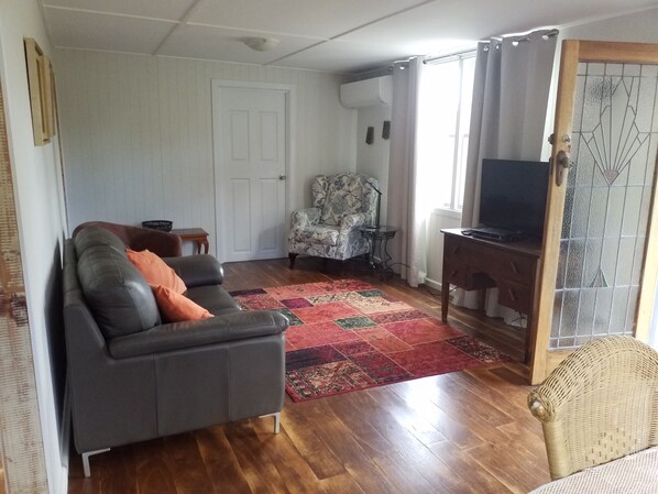 A living room with a dark leather sofa, a patterned armchair, a wooden desk with a television, and a red and green rug.