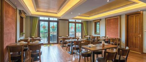 A spacious dining area with multiple tables and chairs, large windows offering a view of the outdoors, and a wooden ceiling with recessed lighting.