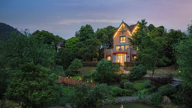 Een groot, twee verdiepingen tellend huis, omringd door weelderige begroeiing en een vijver.