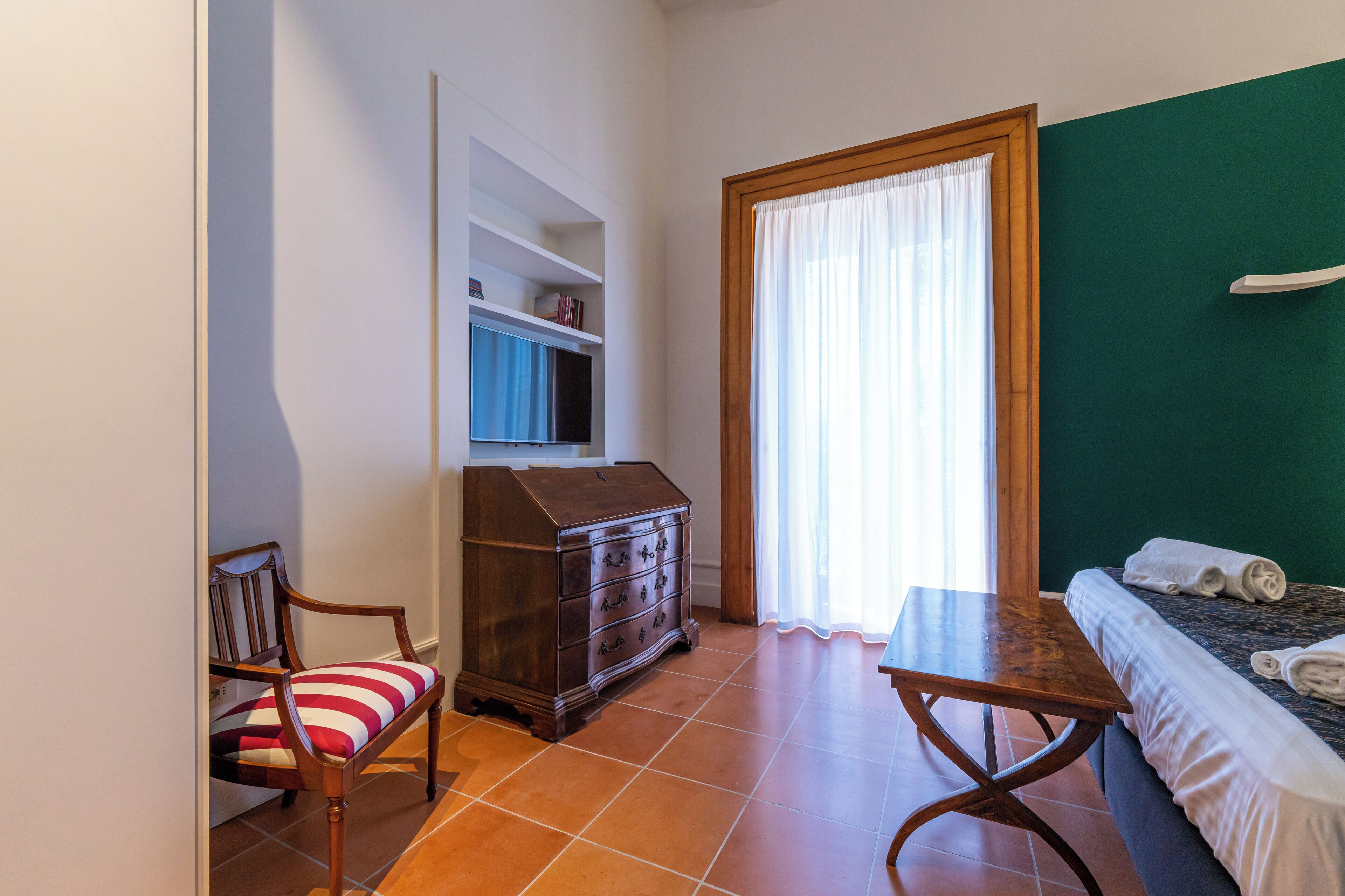 Una habitación con cama, cómoda de madera, silla con cojín a rayas, mesita y ventana con cortinas.