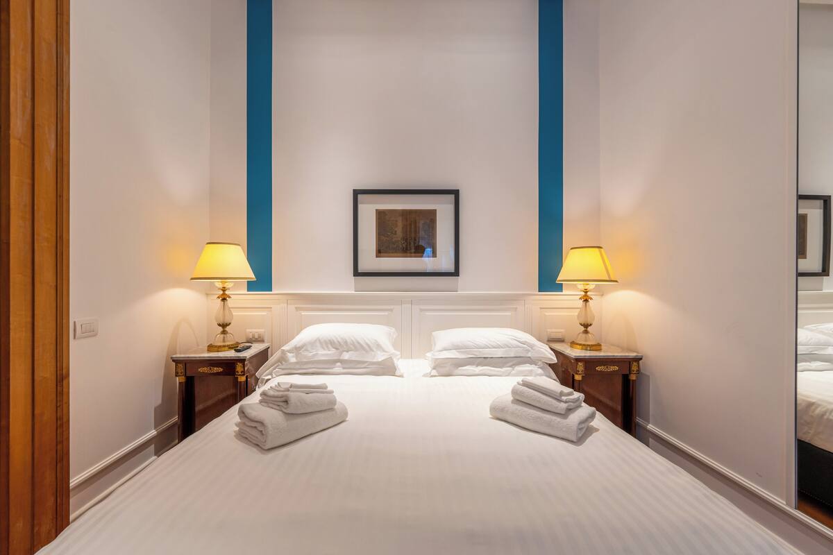 A hotel room with a large bed, two bedside tables with lamps, a framed picture on the wall, and a mirror on the right wall.
