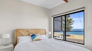 Quarto com uma grande janela com vista para a praia, cabeceira de madeira, colcha branca e abajur.