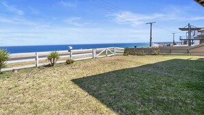 Uma área gramada com cerca branca, uma árvore pequena e vista para o oceano.
