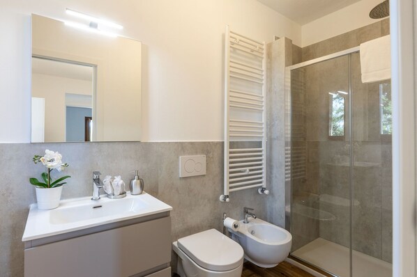 Un baño moderno con un lavabo, inodoro y bidé blancos, cabina de ducha de cristal y un perchero para toallas.