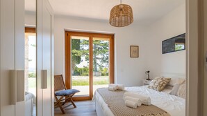 Une chambre avec un grand lit, une chaise en bois, une table de chevet avec une lampe, et une vue sur l’extérieur grâce à une porte vitrée.