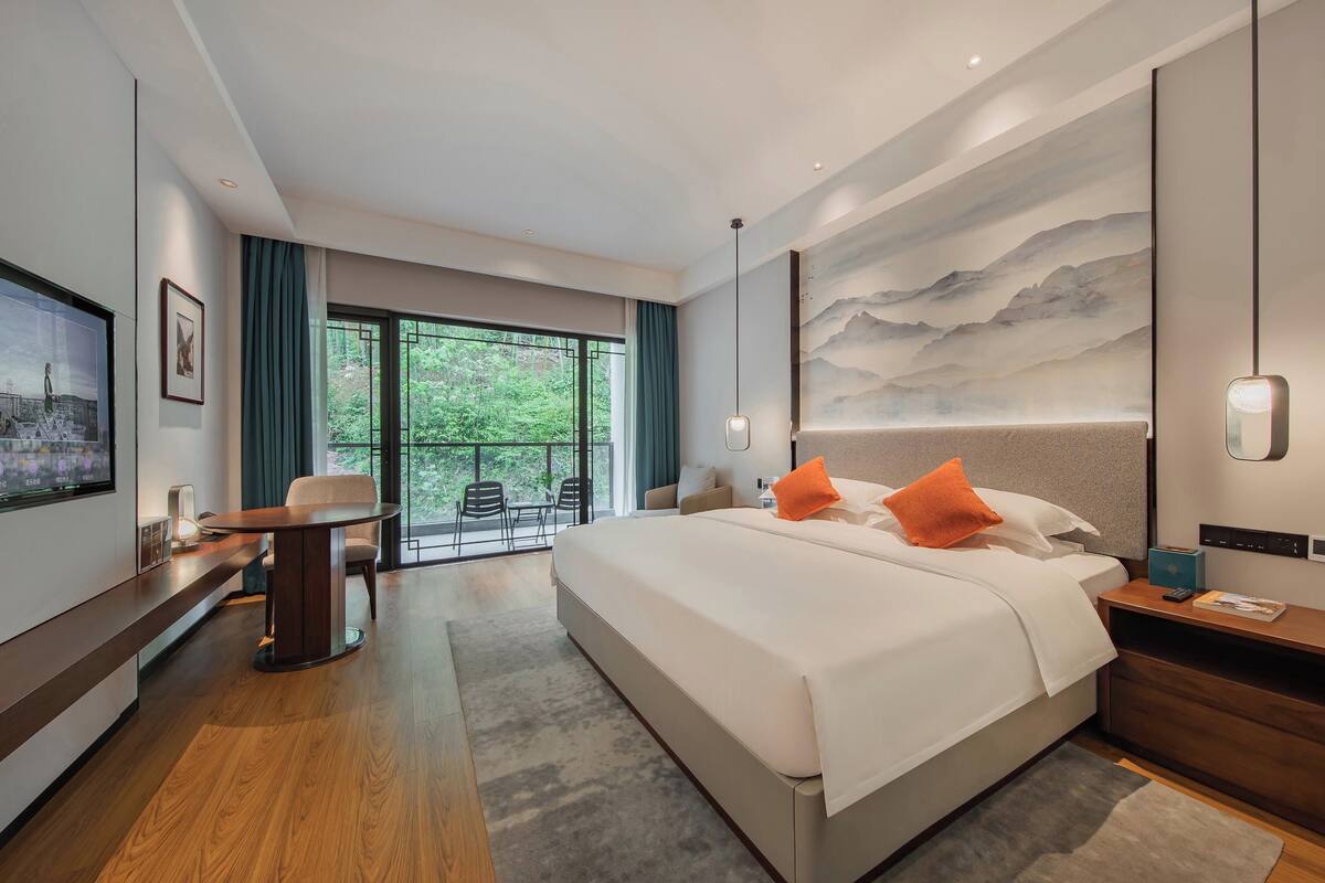 A modern hotel room with a large bed, a desk, and a balcony with chairs.