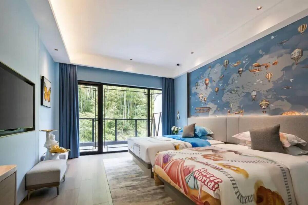 A modern bedroom with a large bed, a smaller bed, a desk, and a bench. There is a large window with a view of greenery.