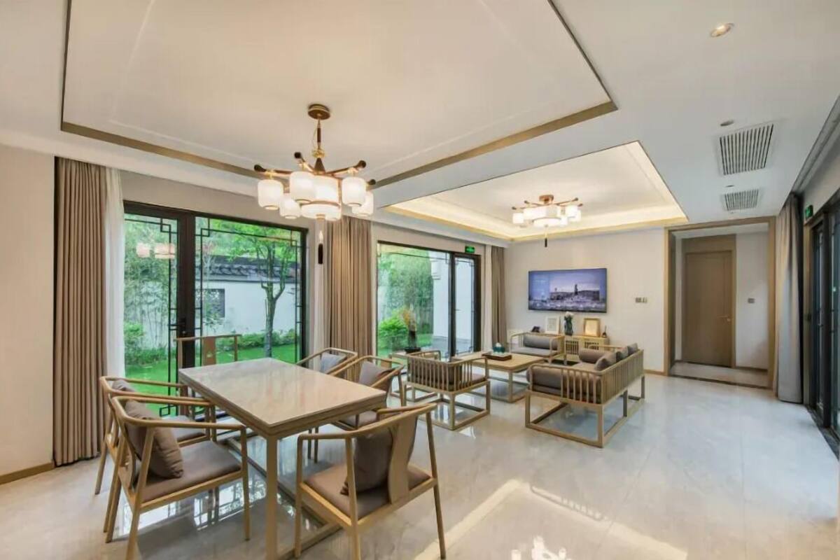 A modern hotel room with a dining area, a sofa set, and large windows offering a view of greenery.